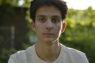 A guy with a white shirt looking right in to the camera. Garden on background