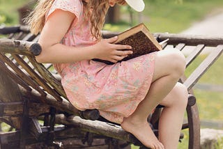 Image of a young girl wearing a pink dress and a white, floppy hat reading a book, sitting on a wooden wheelbarrow