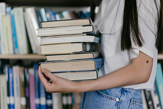 A woman carries a stack of books. She wears a white shirt and jeans.