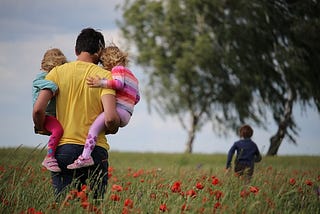 A Dad carrying 2 children with one child running ahead in a field.