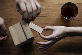 A photo of two pairs of hands playing a game of Jenga. One person is pulling a block out of the wooden tower, which is shot from above. A glass of wine is visible in the top right corner.