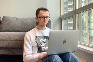 A man in his mid 20s wearing a white sweather and blue jeans. He’s sitting on the floor against a couch. He’s resting a MacBook against his lap. He has black hair and glassses. On the right-hand side of the frame is a window.