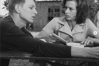 Screenshot from Ingmar Bergman’s Through A Glass Darkly. A young man and woman sit at a picnic table, the woman holds a book in her hands. Subtitle reads: You in your cage, I in mine.