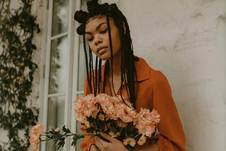 A woman with Box braids leans up against the wall, holding a bouquet of flowers.