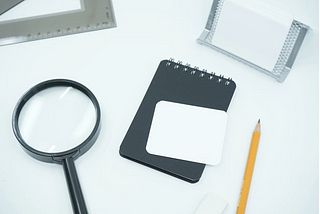 A black notebook on a white table. There is a magnifying glass to its left and a pencil to its right.