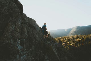 A man standing on cliff face with t-shirt and a hat, looking over a wide forest and mountain range in the background