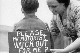 A mother attaches a sign to her son getting ready to ride his bicycle. It reads: ‘Please Mr Motorist. Watch out for me?’