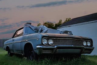An old, dusty car sits abandoned on overgrown grass next to a white building at dusk. The car is covered with a weathered tarp, its paint faded and tires flat. The sky behind shows the soft colors of sunset with scattered clouds, adding a calm, nostalgic mood to the scene.