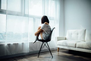 A girl curled up into a ball, sitting on a chair while looking out the window.