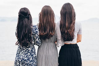 Three slim brunette women stand close with their backs to the camera, looking towards a misty landscape. Two wear long flowing dresses, the other a long black skirt and tight white top.