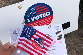 A hand holding a small American flag with an “I Voted” sticker and an envelope marked to indicate it is a mail-in ballot. Backdrop is a larger piece of cardboard with a larger “I Voted” sticker.