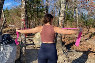 a woman performing resistance band exercises with a pink resistance band outdoors at a campsite