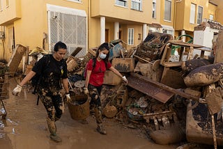 Two women wearing boots full of mud and walking on a pavement all brown due to the mud are carrying one bucket (one from each handle, with the bucket in between them). Behind them, very close, is a big pile of mud-covered debris. In the background, the houses of people in light sand-colour rendered walls.