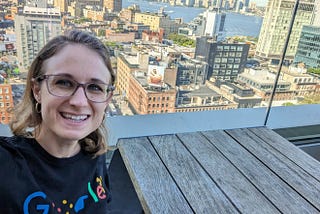 Lisie wearing a Google accessibility shirt, with a beautiful view of Manhattan behind her. The shirt says Google, but with the ‘o’s written in braille, the second ‘g’ represented in sign language, and the ‘e’ shown with a person sitting on it as if the letter were a wheelchair