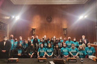 Members of the group Long Covid Moonshot and other advocates and researchers pose in a wood-paneled government room, wearing teal T-shirts advertising the need for moonshot-level funding.