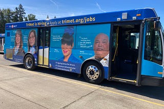 A Gillig 30 foot bus operated by TriMet wrapped with an advertisement about job opportunities at TriMet. The ad features four TriMet employees talking about how much they enjoy working at the transit agency. Cheree, a bus trainer says, “The benefits are awesome, the pay is outstanding.” Caryn, a planning engineer says, “Helping to build the future of transit is rewarding.”