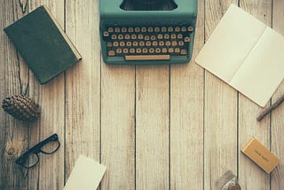 A teal typewriter on a white wooden floor, surrounded by glasses, books, and other writerly things. Photo by Dustin Lee.