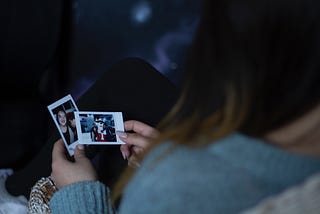 The image shows the back of a woman’s head. From over her shoulder, you can see she is holding two polaroid photos.