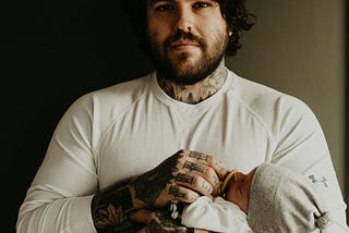 An adult man with a beard and tattoos holds a pacifier to an infant’s mouth.