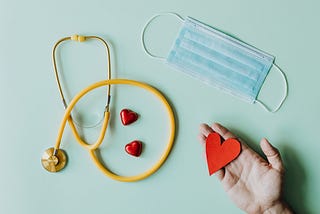 A yellow stethoscope, disposable mask, and two ceramic hearts are laid out on a turquoise background. A hand hovers over all of them, holding a red paper heart in its palm.
