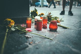 Candle and flowers on the ground in a public street as people walk by and one person stands by them