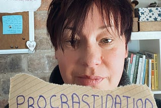 An image of a woman with short hair holding up a piece of paper that reads ‘procrastination’.