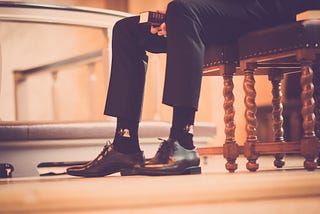 A picture of a man’s legs, he’s wearing a suit, sitting on a pew in a church holding a bible. (He’s supposed to represent a pastor).