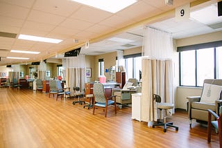 Five consecutive beige hospital chairs are separated by cream curtains pulled back with one extra seat for each station and a rolling charting cart.