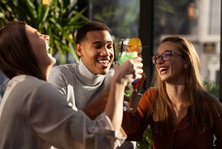 Three adults sitting around the same table in a bar, clicking their drinks together with large smiles on their faces. They are clearly having a great time together.