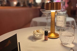 A table covered with a white mantel, in the center a gold lamp, a small pot with sugar sachets and a personalized hand sanitizer. a glass full of sparkling water to the right and to the left a black and gold menu.