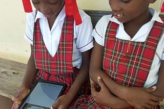 Two girls in school uniform sit outside on a wooden bench. One holds a tablet and the other has her arms crossed. Both are looking at the tablet.