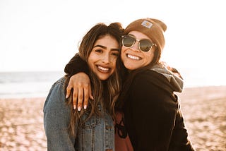 Two young women, best friends, embracing on the beach, one with her arm around the shoulder of the other, the two of them, heads touching, smiling, looking out at the camera.