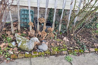 Weathered stone goose garden statue lying on its side in a leaf-covered flower bed bordered by old bricks