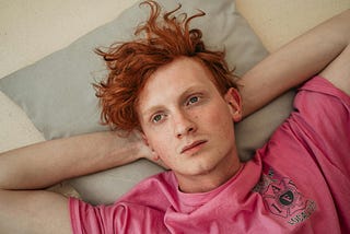 Young man lying on his back looking up, red hair pink shirt