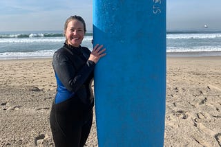 Jeanie stands on the beach in a wet suit, holding a surf board.