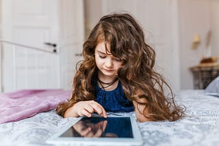 A young girl with long curly hair lying on bed using a tablet.