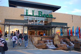 Entrance to LL Bean store with kayaks outside and large glass windows. There is a large sign in green letters that says LL Bean and many people entering the store.