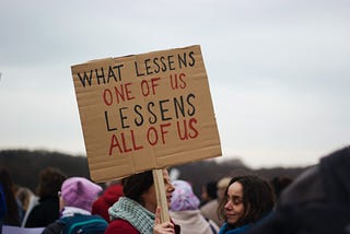 Woman holding a sign that reads: “What lessens one of us lessens all of us.”