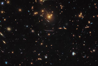 Dieses Bild des Hubble-Weltraumteleskops der NASA/ESA zeigt eine bunte Mischung verschiedener Galaxien, von denen einige zu derselben größeren Struktur gehören. Der Hintergrund ist schwarz mit vielen Streifen und Sprenkeln aus weißem, gelbem und hellblauem Licht. Einige sind kleine Flecken, andere ziemlich groß.