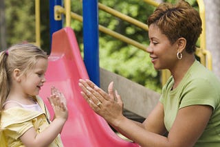 White child with long blond hair/pigtails playing paddycake with black women with short hair and green shirt on playground