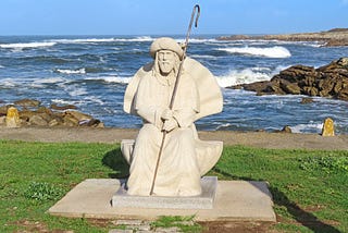 White statue of Saint James holding an iron staff. Statue is sitting on green grass. Ocean waves crash over rocks in the background.