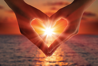 Two hands making heart shape with ocean in background and sunset shining through the centre