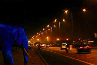 An ultramarine elephant watches cars go by on a busy highway at night, bathed in orange light