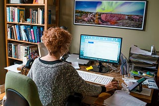A woman with short red hair sits in a home office at a computer in front of a bookcase.
