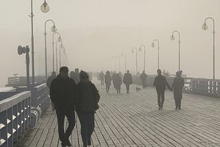 Silhouettes of people walking along a wooden pier in mist with street lamps on each side.