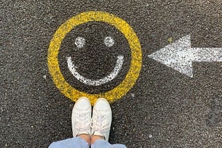 A smiley face in chalk on the ground beneath a woman’s feet, with an arrow pointing at it.