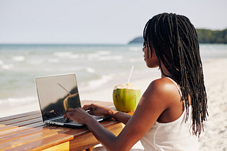 Black woman with long, natural locked hairstyle, typing on a laptop at the beach, drinking from a fresh coconut