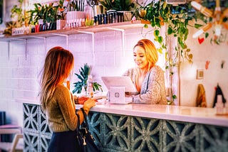 A woman at a shoppe and a Receptionist at a computer at the counter. Many plants on a high shelf
