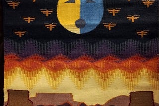 A pictorial weaving by Bobbi Joe Whitehair titled “Moon of the Dragonflies.” It shows a yellow and blue moon on a black background  over a desert landscape. There are dragonflies on each side of the moon.