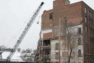 A wrecking ball on a crane swinging towards an old partially-wrecked brick building.